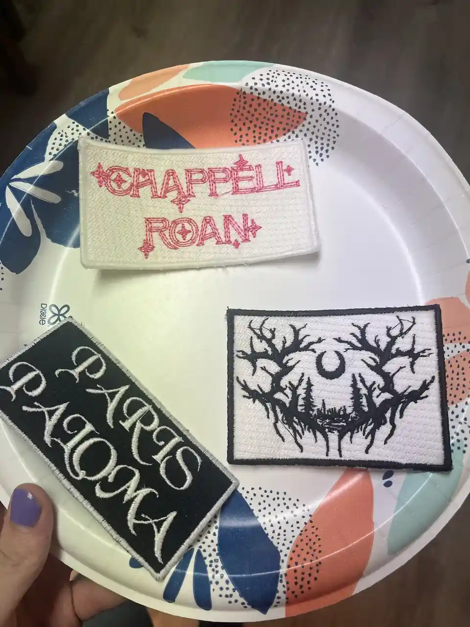A hand holds a patterned paper plate displaying three embroidered patches: one white with red 'CHAPPELL ROAN' text, one…
