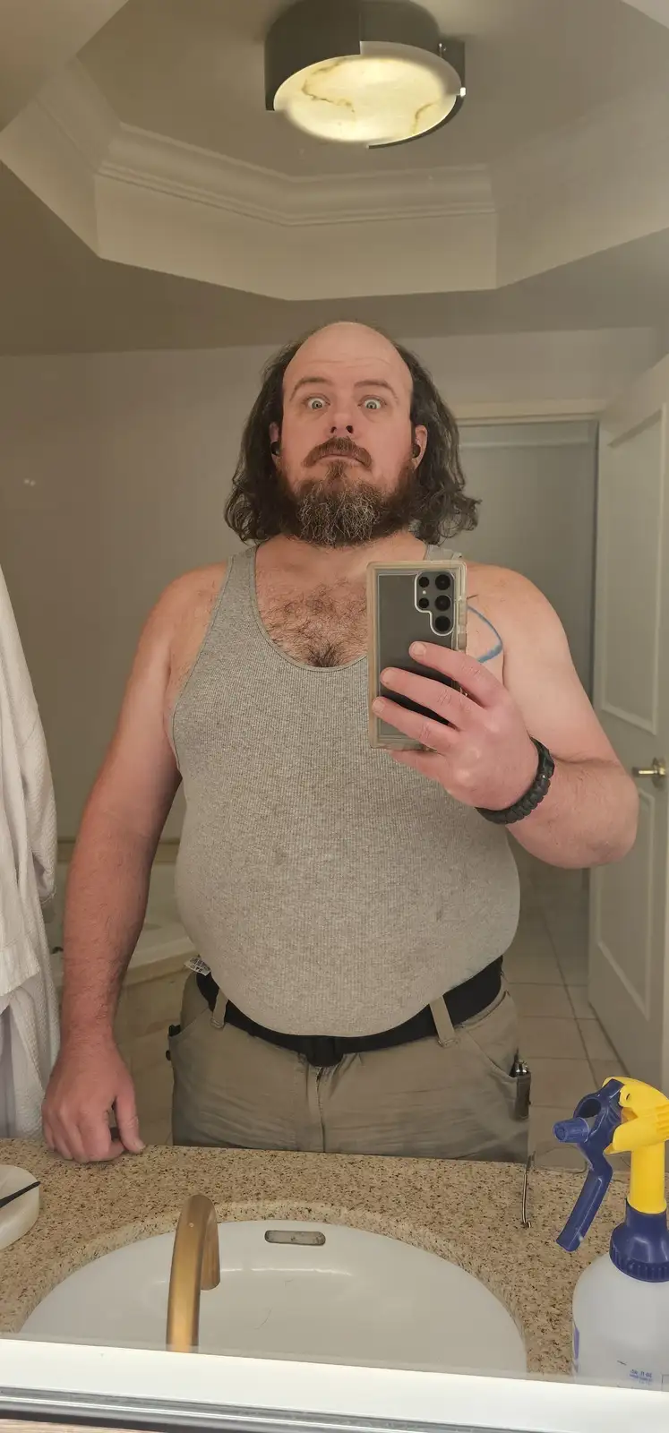 A man with a beard and long hair takes a mirror selfie in a bathroom, looking shocked or horrified while holding a…
