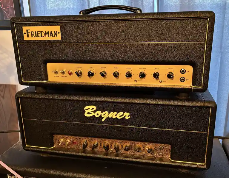 Two stacked guitar amplifiers, a Friedman head on top of a Bogner head, both with black textured tolex and gold trim. The…