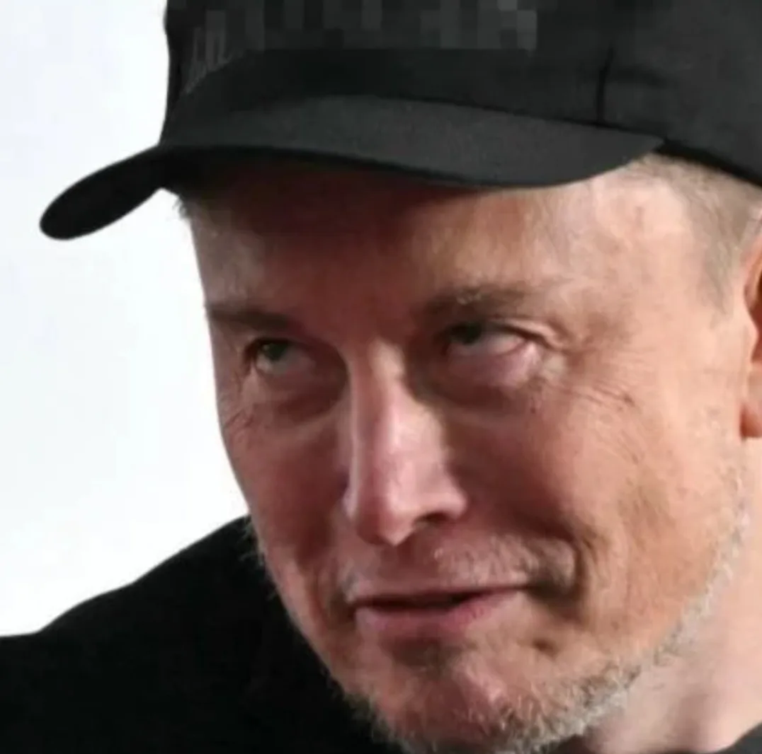 A close-up photo of Elon Musk wearing a black baseball cap, looking sideways with a subtle smirk. The image has pixelated…