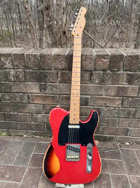 A red and black electric guitar with a worn, sunburst finish leans against a brick wall outdoors. The guitar's body shows…