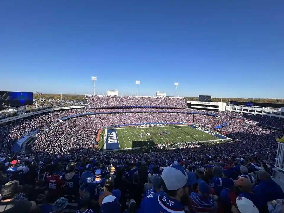 A wide-angle photo of a packed Buffalo Bills football stadium under a clear blue sky, showing fans in blue and red jerseys…