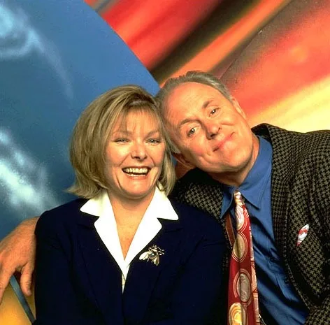A man and woman are posing together in front of a colorful abstract background. The man has his arm around the woman and is…