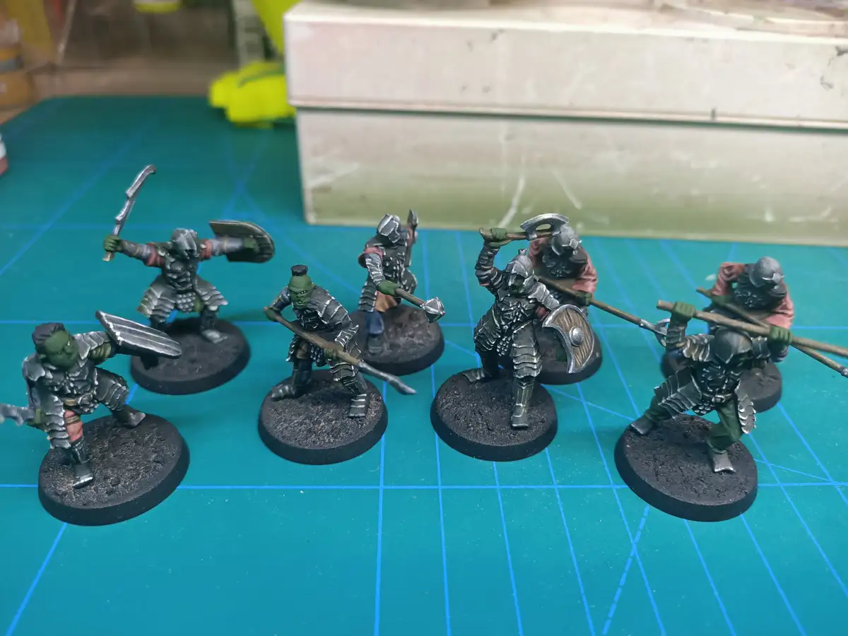 A photo shows six painted miniature orcs in silver armor standing on black circular bases on a green grid mat. Each orc…