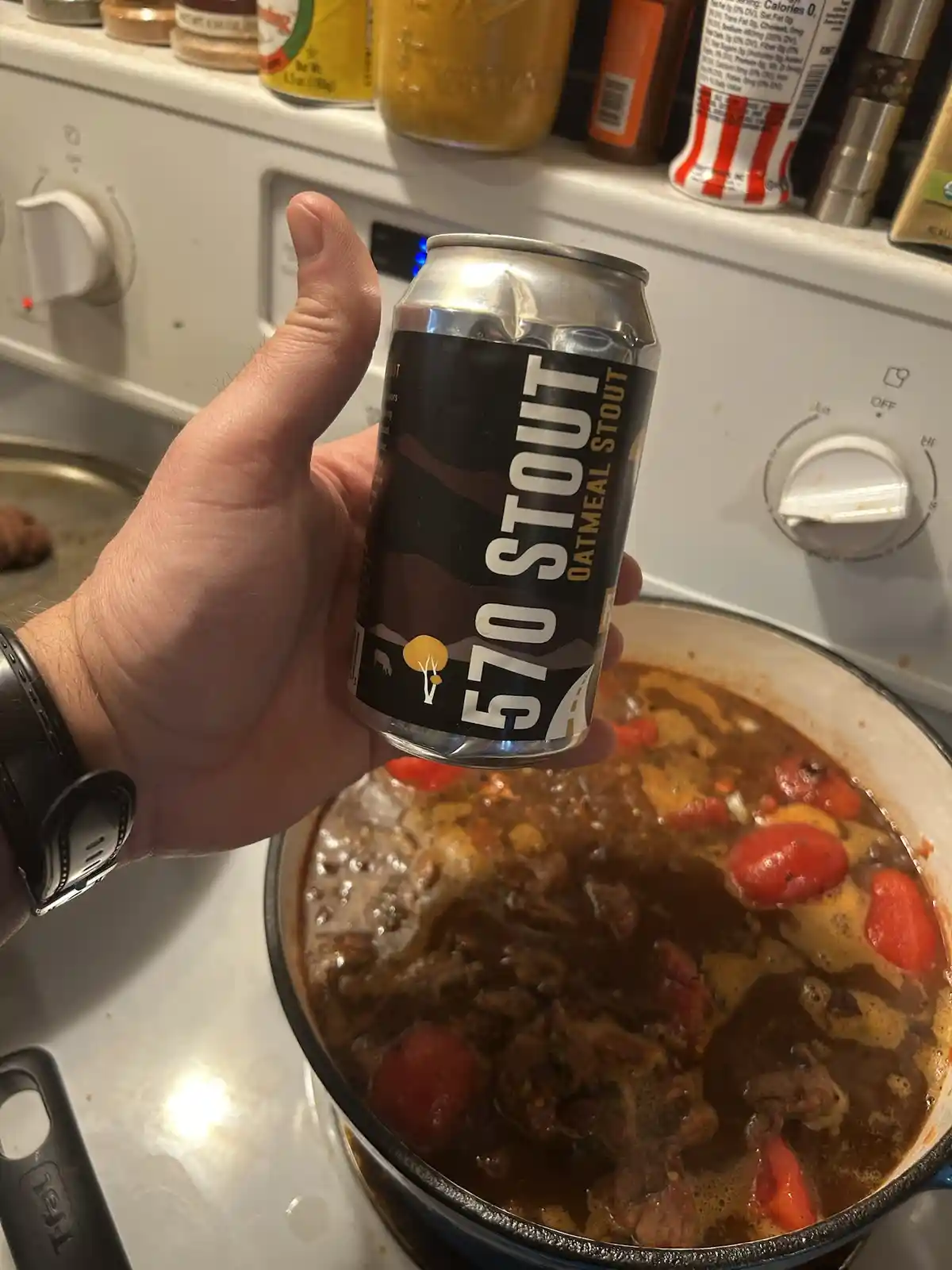 A hand holds a can of '570 Stout Oatmeal Stout' beer directly over a pot of simmering stew on a stovetop, with kitchen…