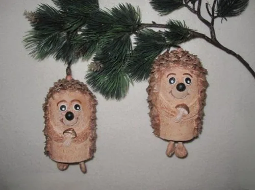 Two pinecone ornaments shaped like hedgehogs with googly eyes, black noses, and tiny feet hang from a pine branch against a…