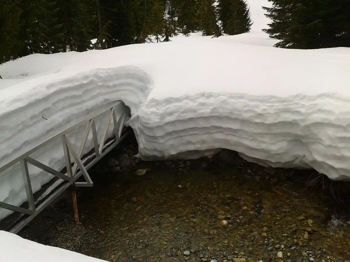 A metal footbridge is partially buried under a thick, layered snowdrift that has formed a natural arch over a shallow, rocky…
