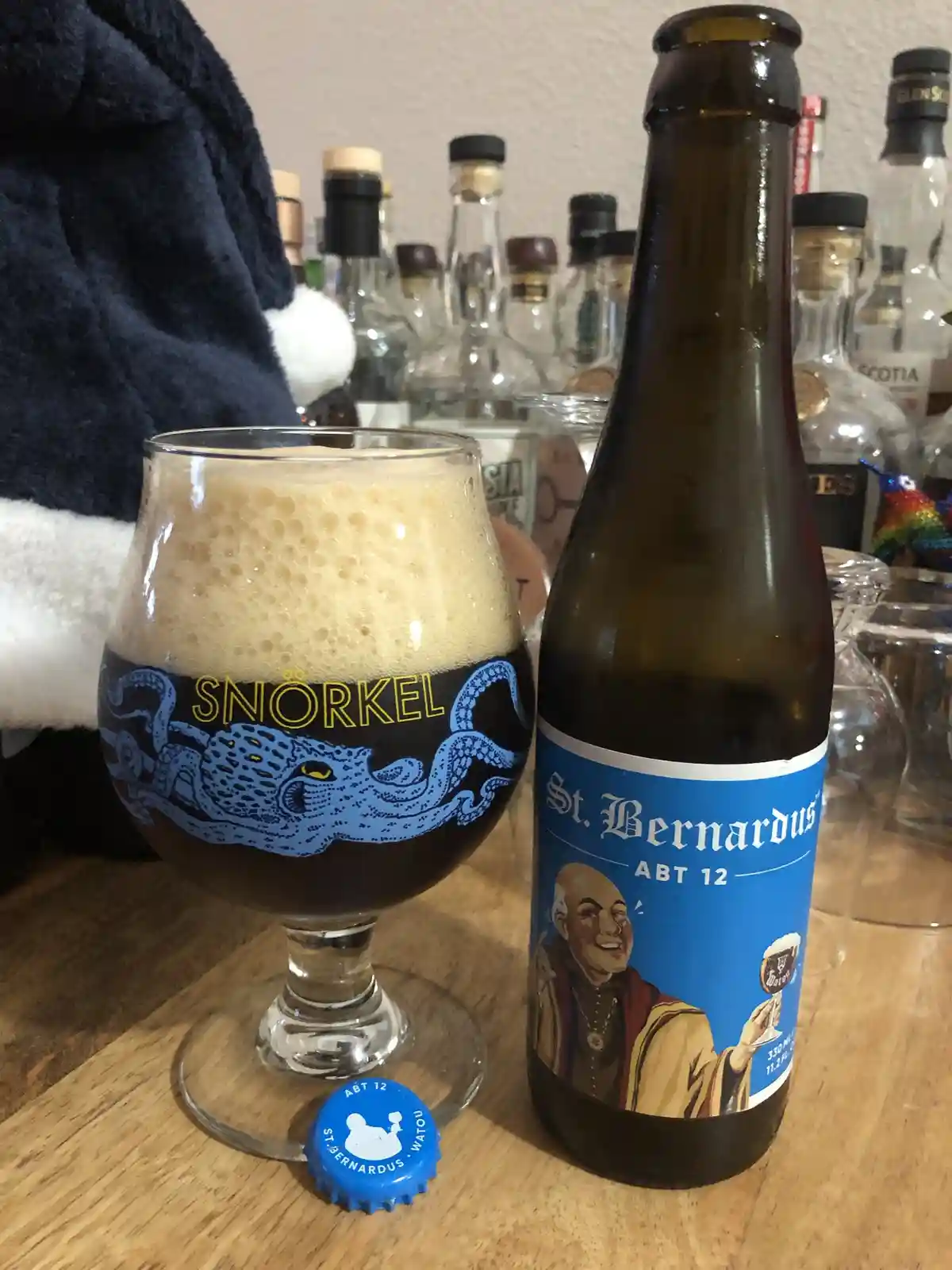A glass of dark beer with a thick, foamy head sits next to a St. Bernardus ABT 12 bottle on a wooden table. The glass is…
