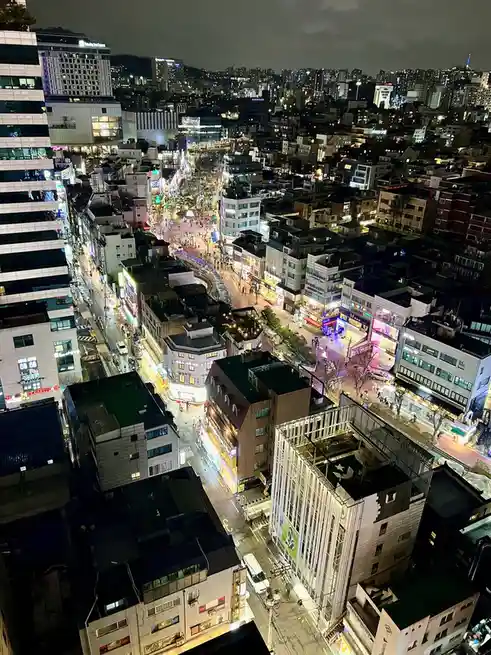 An aerial night photo of a densely packed urban area with glowing streets, numerous buildings, and visible traffic and…