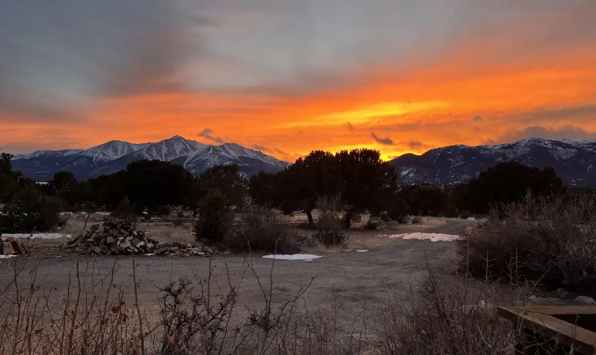 A scenic photo of a mountain range at sunset, with orange and yellow clouds above snow-capped peaks, a gravel path, trees…