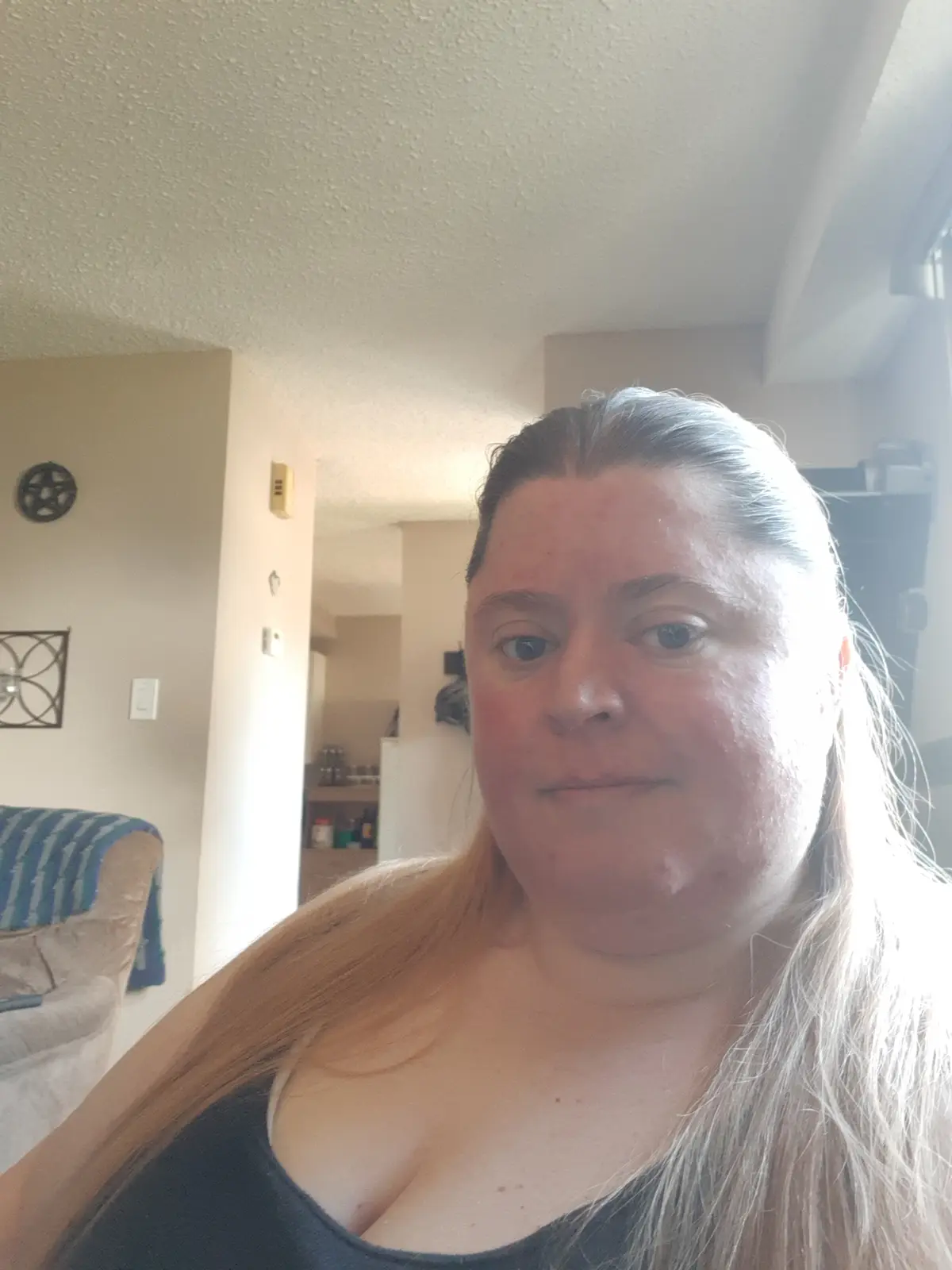 A person with long blonde hair and a black tank top looks directly at the camera with a neutral expression in a brightly lit…