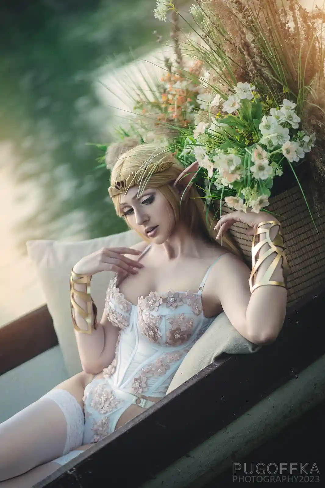 A person in an elaborate fantasy cosplay, resembling a female elf with pointed ears, golden headpiece, and ornate white and…