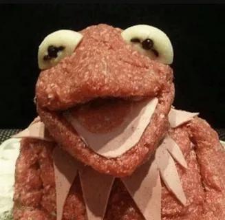 A close-up image of Kermit the Frog, whose entire body is crafted from ground meat, with white cheese slices forming his…