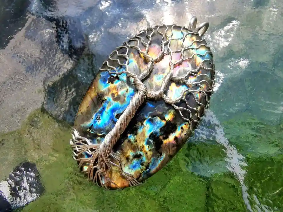 A handcrafted silver wire tree of life pendant rests on a vibrant, iridescent abalone shell, which is floating on the…