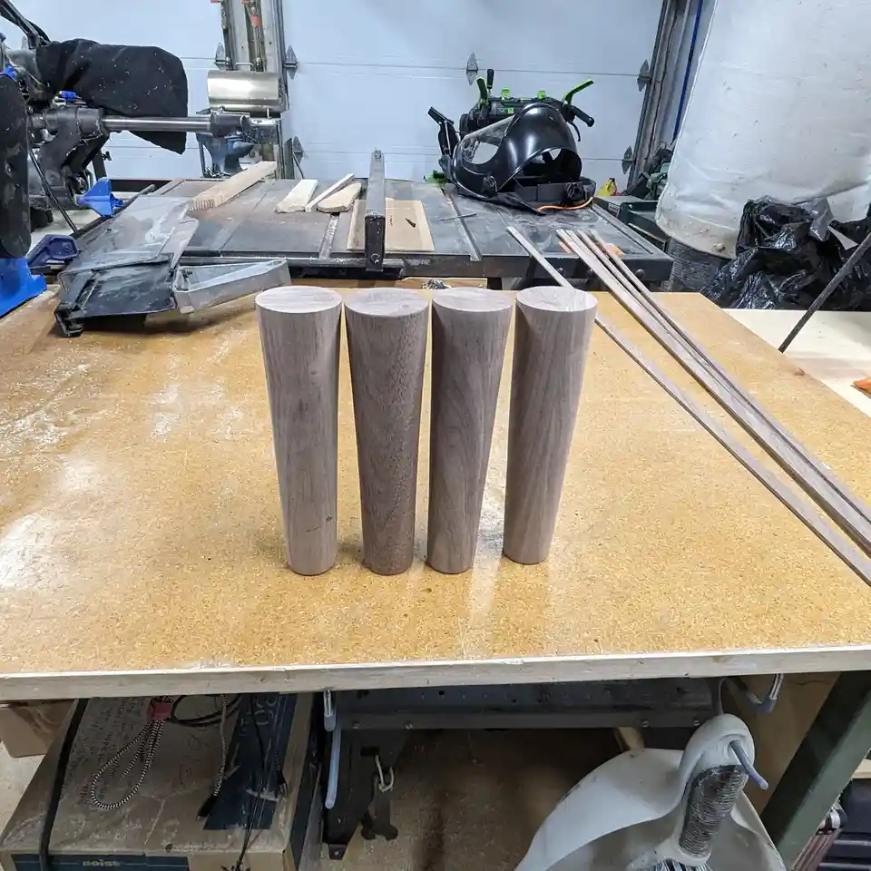 Four freshly turned wooden dowels stand upright in a row on a workshop workbench, surrounded by woodworking tools like a…