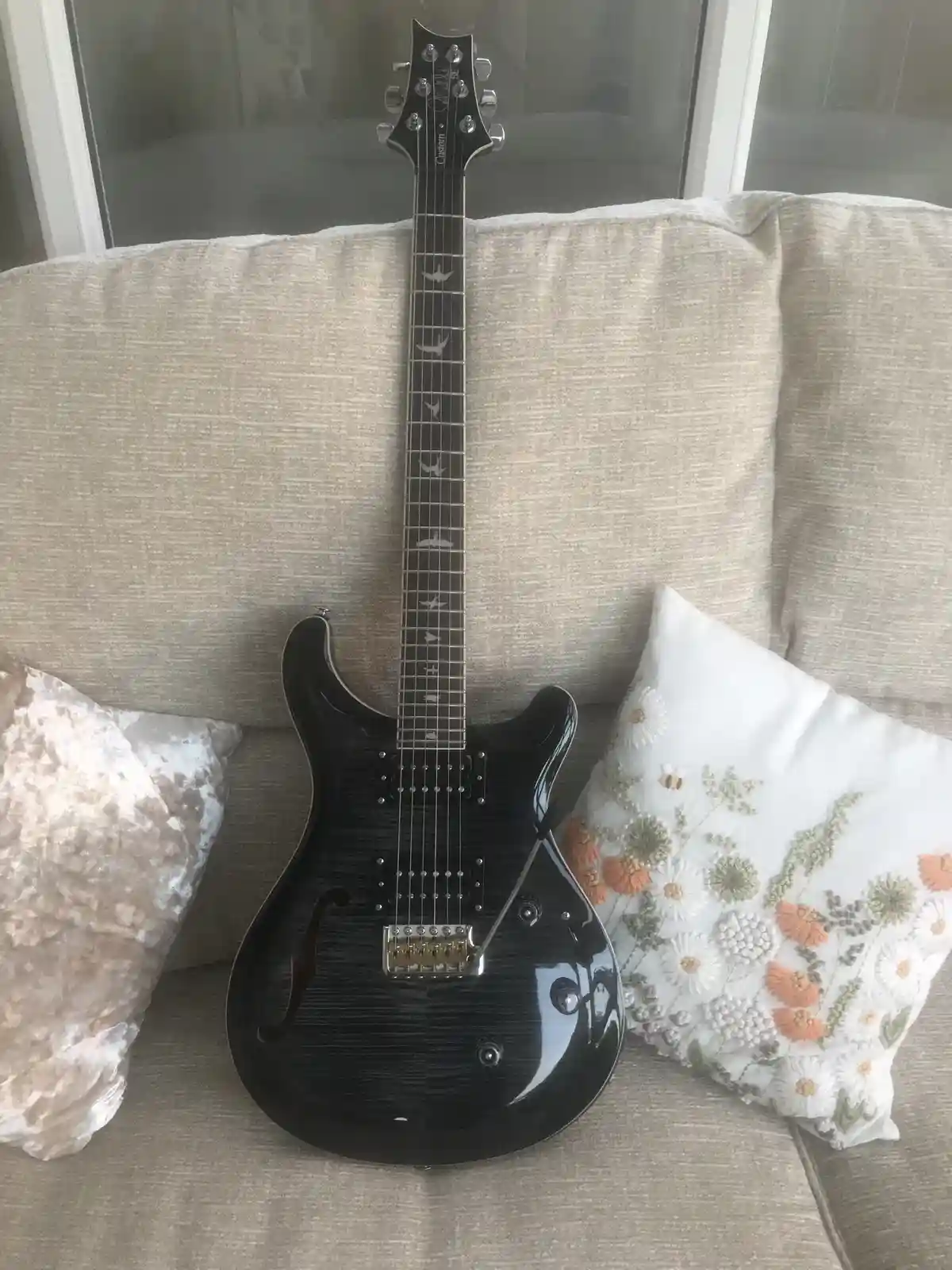 A black electric guitar with bird inlays on the fretboard is propped upright on a beige couch, flanked by two decorative…