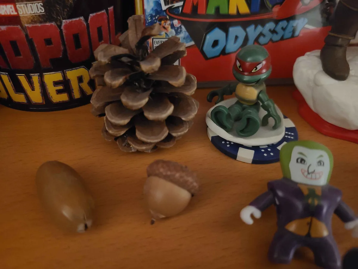 A wooden surface displays a pinecone, an acorn, a green ninja turtle figurine, and a joker figurine, with Marvel Studios and…