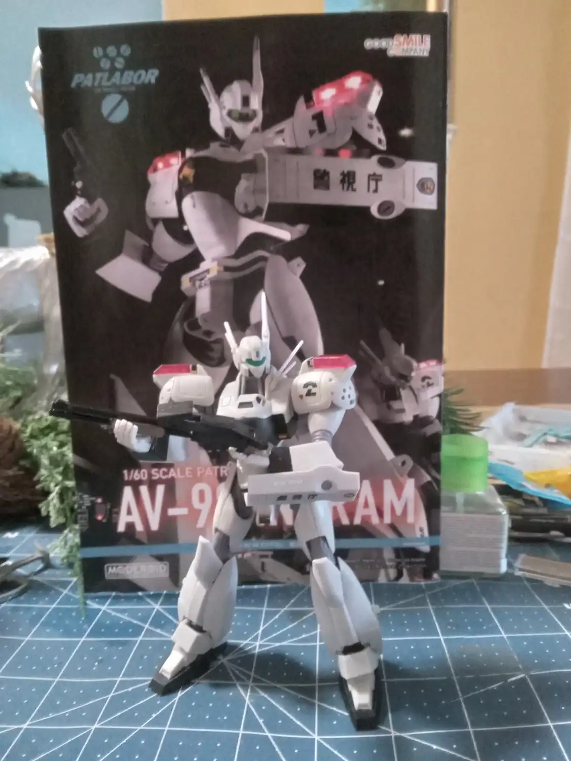 A white and grey 1/60 scale Patlabor AV-98 model robot stands on a blue grid mat in front of its box, holding a black rifle…