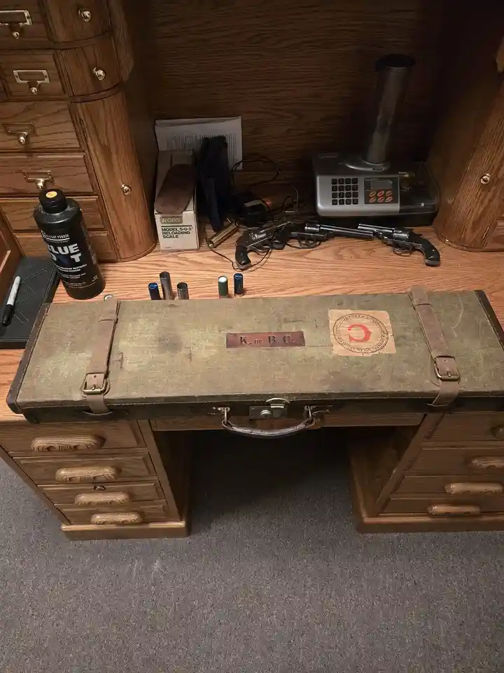 A vintage olive-green canvas toolbox with leather straps and a metal handle sits on a wooden desk, labeled 'K. OF B.C.' with…