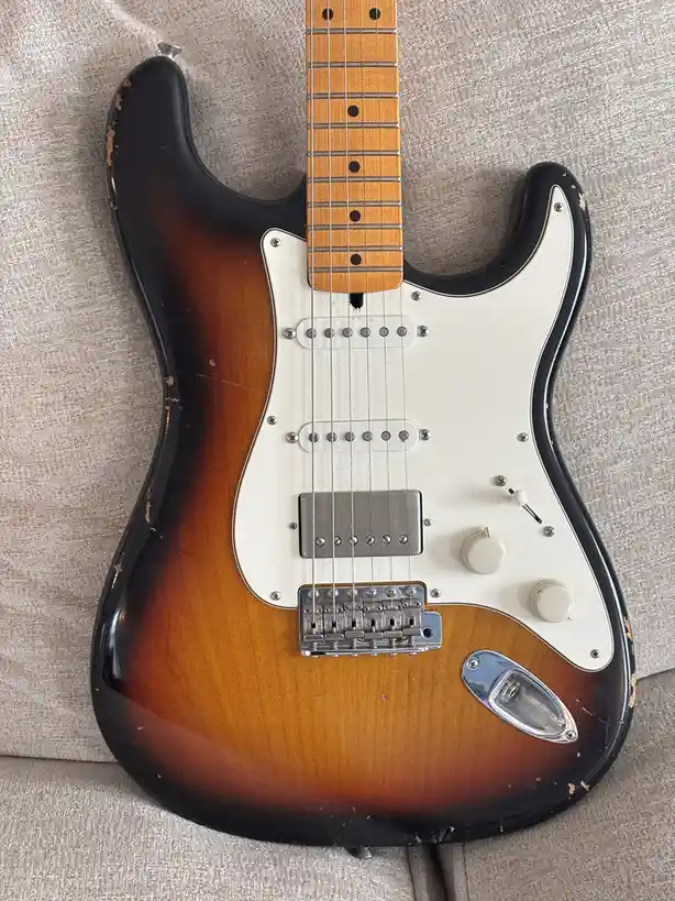 A sunburst-finish electric guitar with a worn, chipped black edge and white pickguard rests on a beige fabric couch. The…
