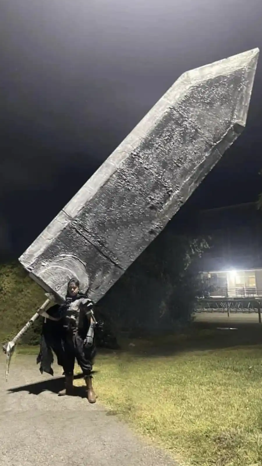 A person in dark fantasy armor stands outdoors at night, holding an enormous, crudely crafted sword that dwarfs them. The…