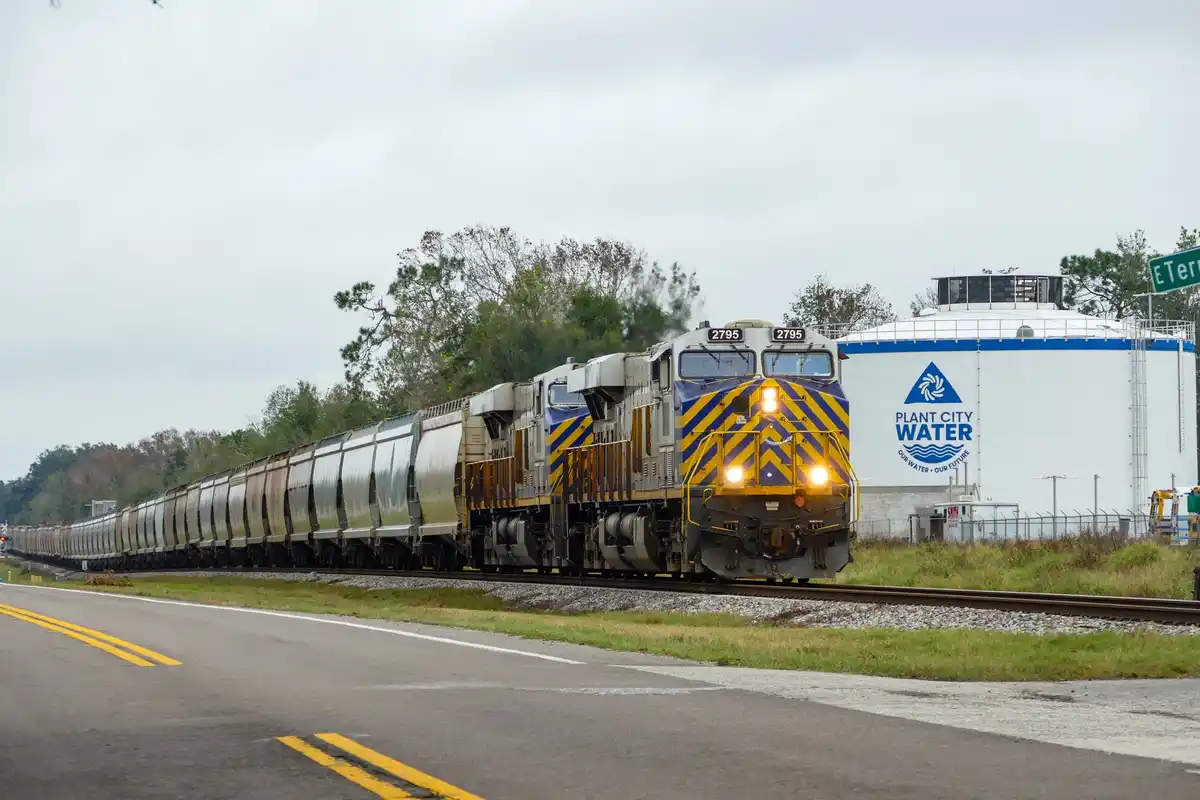 A long freight train with locomotive number 2795 travels past a large white water tank labeled 'PLANT CITY WATER' under an…