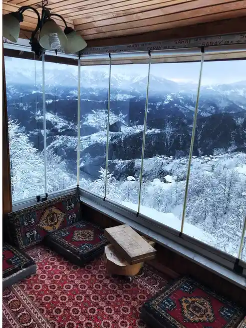 A cozy room with patterned rugs and cushions sits before a large window overlooking a snowy mountain landscape. The window…