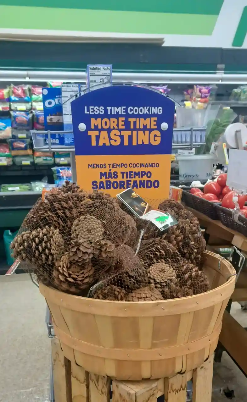 A wooden basket filled with pinecones sits in a grocery store produce aisle, with a blue and orange sign behind it that…