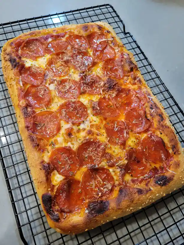 A rectangular pepperoni pizza with golden-brown crust, melted cheese, and crispy pepperoni slices sits on a black wire…