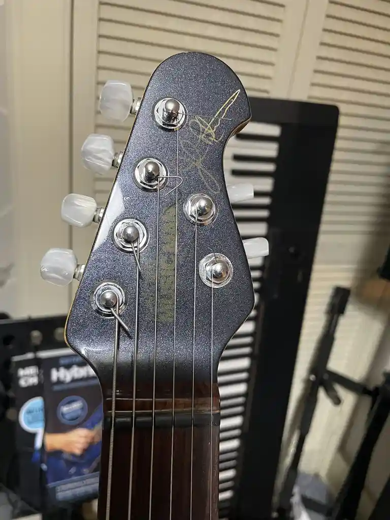 A close-up photo of a guitar headstock with a visible signature in gold ink, likely from a band, overlaid with the text 'i…