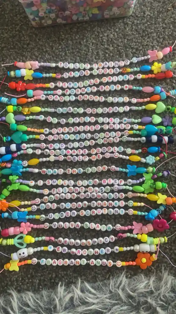 A collection of colorful beaded phone charms laid out on carpet, each charm spelling out a humorous or crude phrase using…