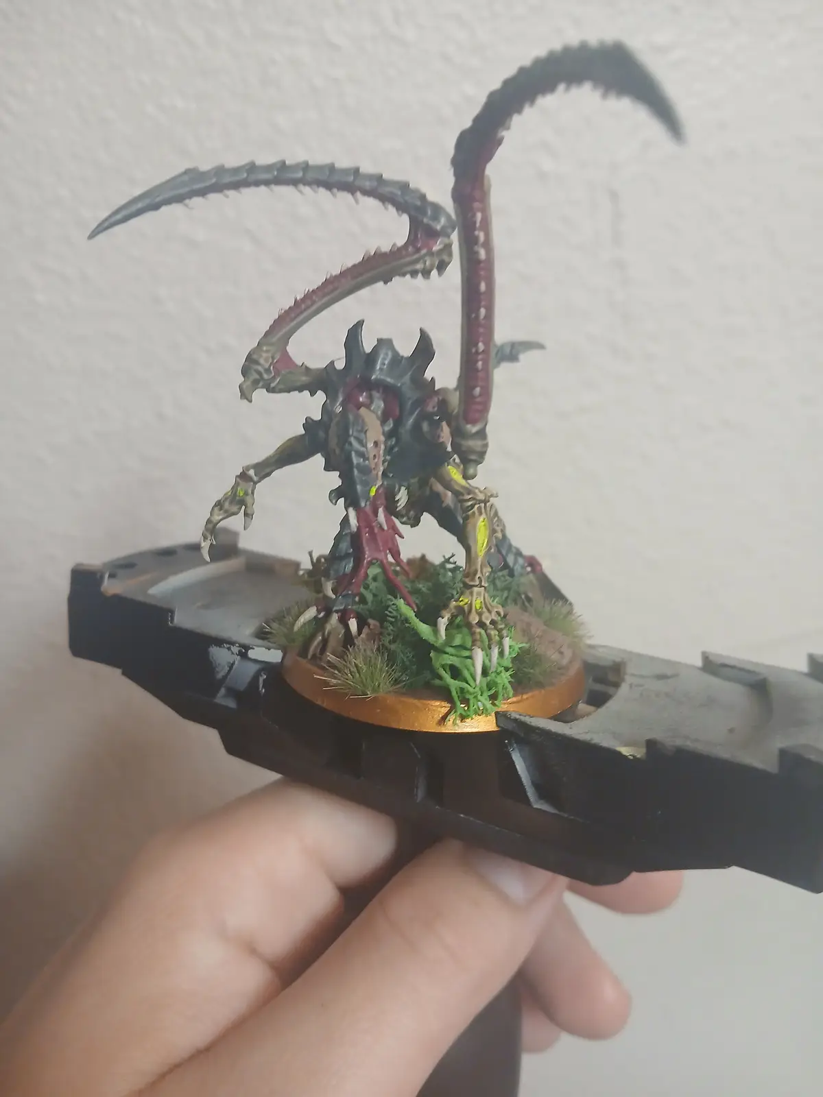 A hand holds a Warhammer 40,000 Tyranid bio-weapon miniature on its base, which is mounted on a black plastic display stand…