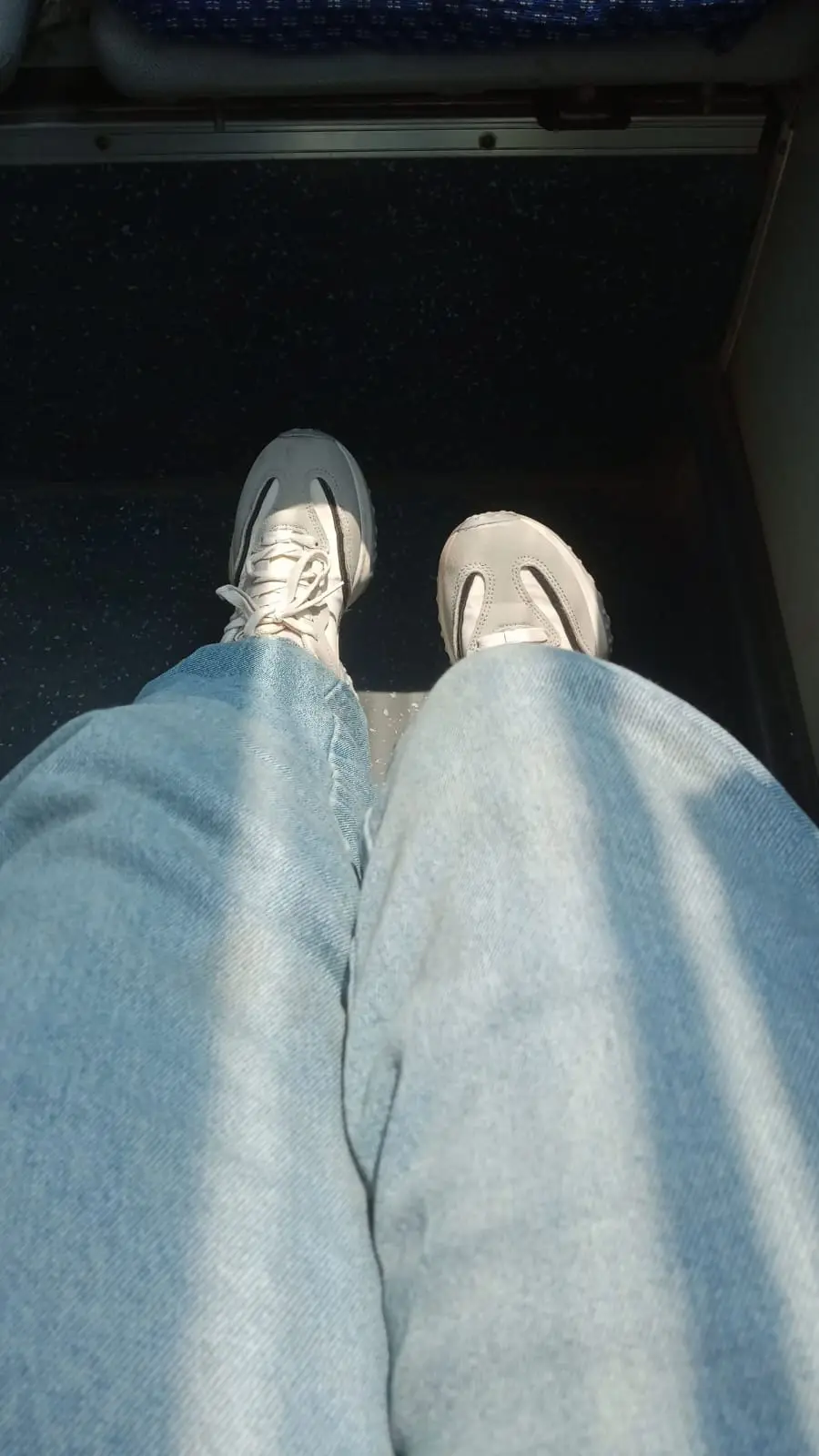 A first-person view from someone sitting on a train or bus, showing their light blue jeans and light gray sneakers on a dark…