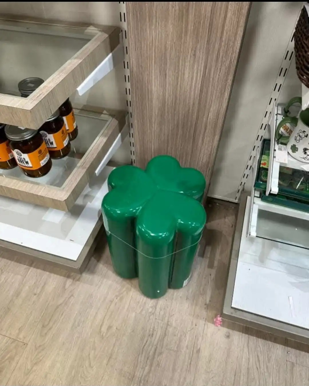A green, four-leaf clover-shaped object sits on a wooden floor in a store aisle, surrounded by shelves with jars and other…
