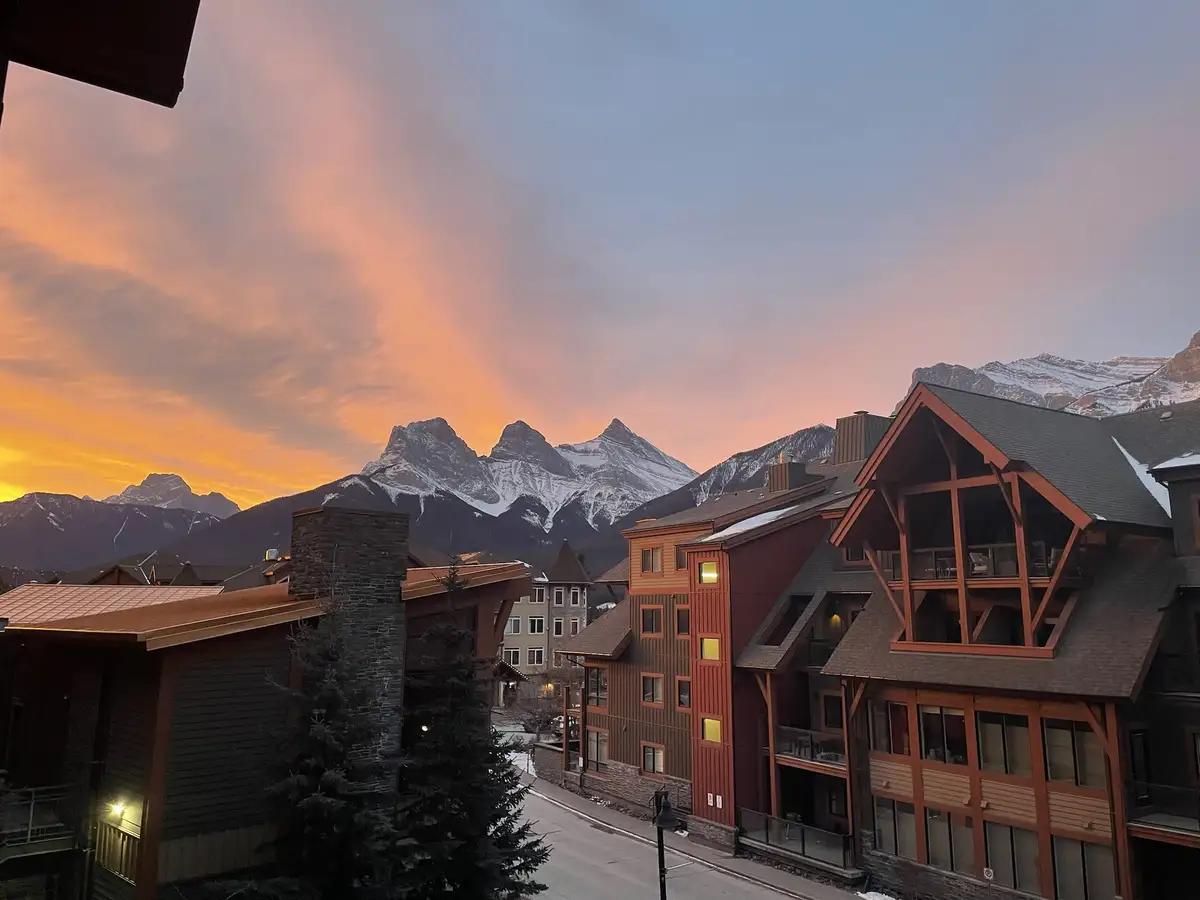 A scenic photo of a mountain town at sunset, featuring snow-capped peaks under a vibrant orange and pink sky, with wooden…