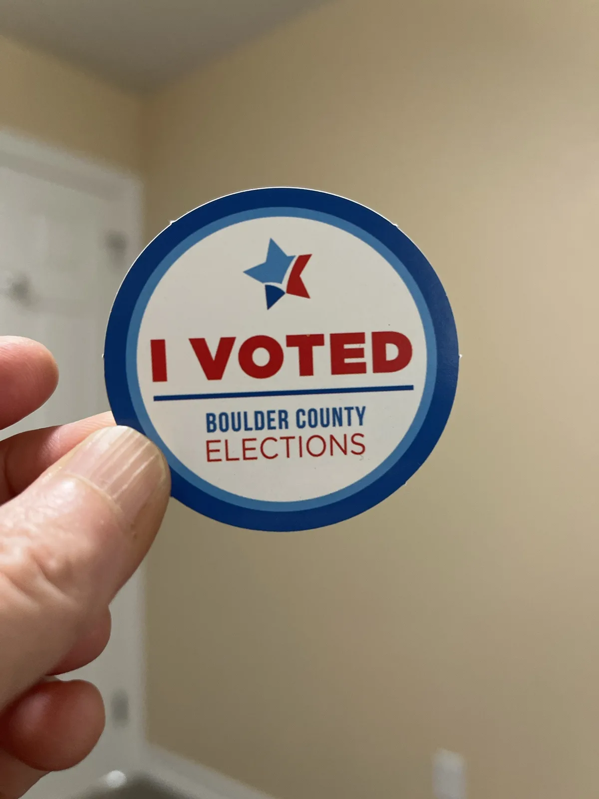 A hand holds up a circular 'I VOTED' sticker from Boulder County Elections, featuring a blue and red star logo above the…