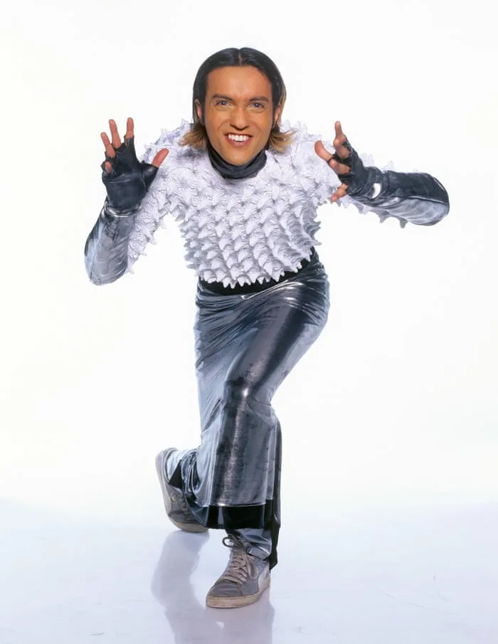 A man with long hair and a wide smile poses energetically against a plain white background, wearing a white spiky-textured…