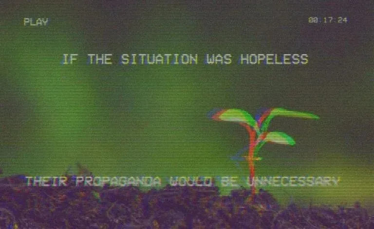 A grainy, VHS-style video frame shows a small, colorful plant sprouting from dark soil against a green background. The text…