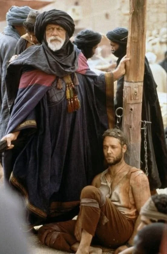 A man in a dark turban and robe stands beside a wooden post with a chain, while a shirtless, chained man sits on the ground…