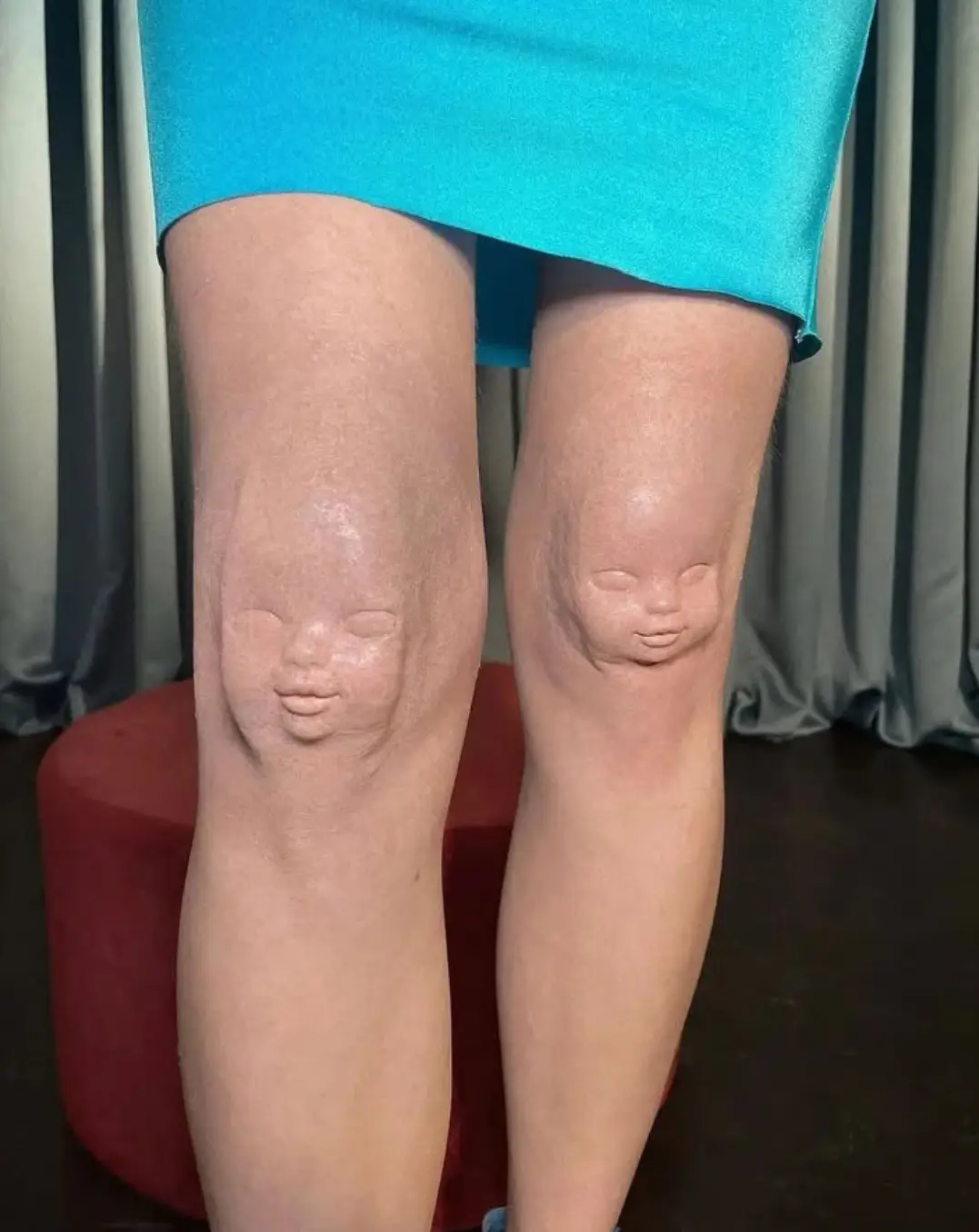 A person wearing a blue skirt stands with their knees prominently displayed, each knee featuring a detailed, realistic…