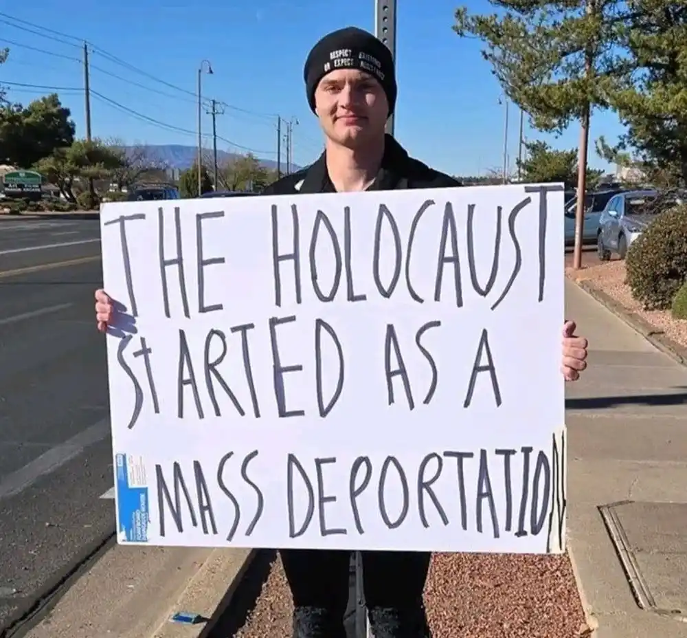 A young man wearing a black beanie stands on a sidewalk holding a large white sign with handwritten text that reads 'THE…
