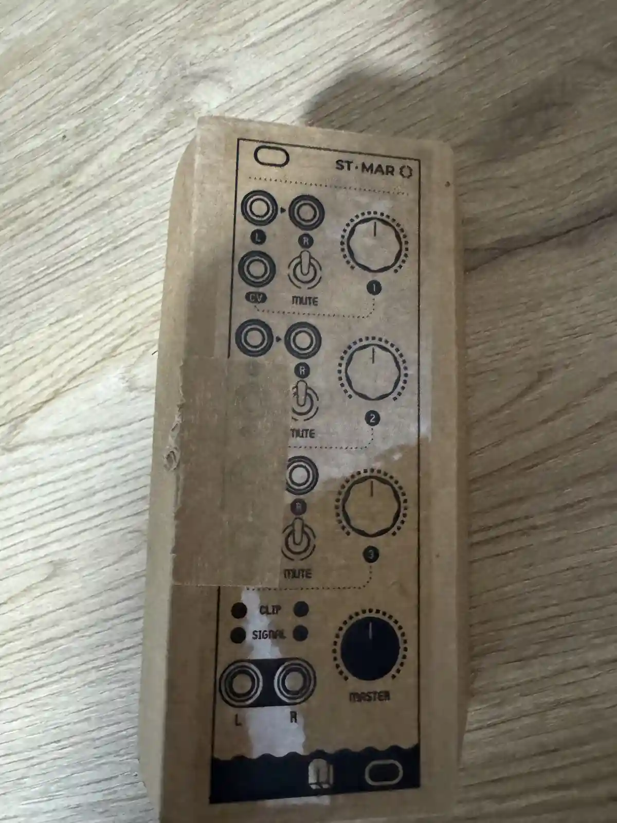 A close-up of a cardboard box with a printed schematic resembling an audio mixer or synthesizer interface, featuring knobs…