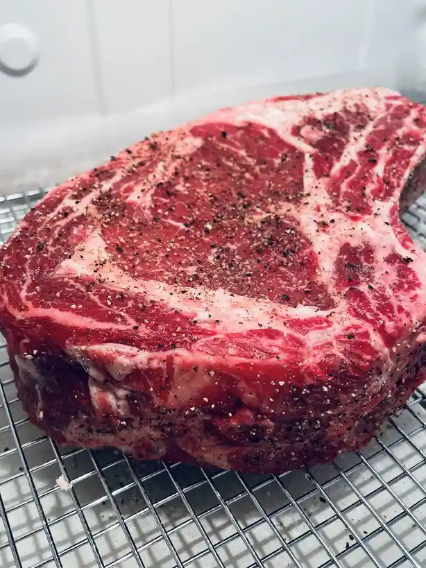 A large, raw, marbled steak seasoned with black pepper and salt sits on a metal cooling rack, with a white tiled background…