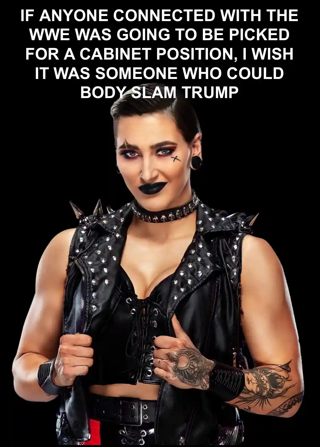 A muscular woman with short dark hair, heavy black makeup, and tattoos on her arms wears a black studded leather vest over a…