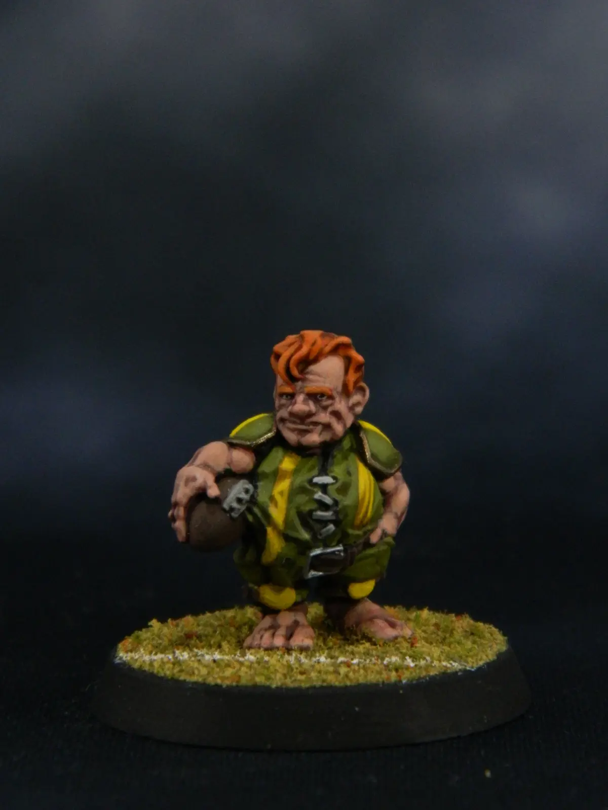 A painted miniature figurine of a stout, orange-haired, barefoot humanoid in a green and yellow outfit, crouching and…