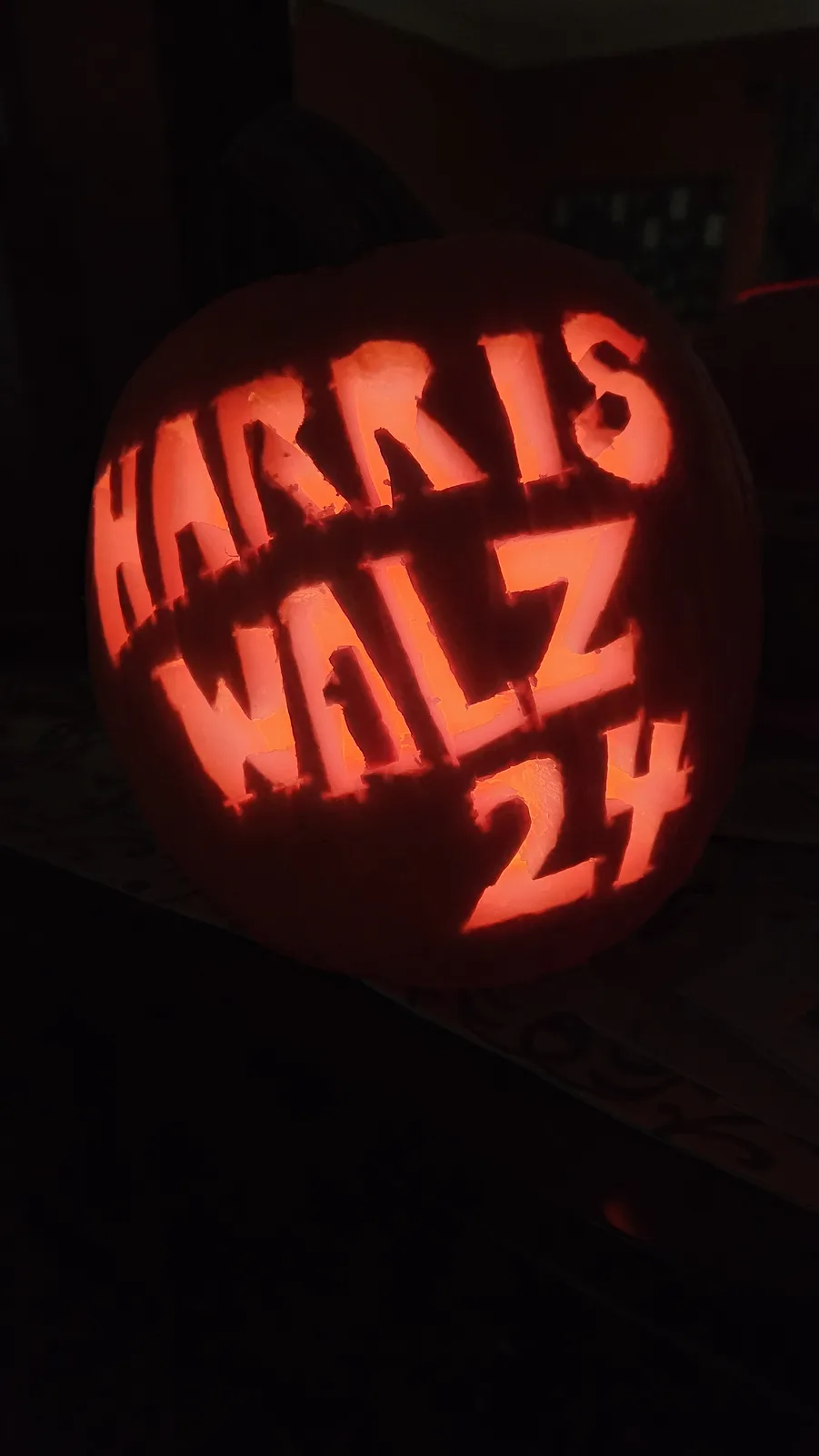 A carved Halloween pumpkin with glowing orange light from within, displaying the text 'HARRIS WALTZ 24' in large, blocky…