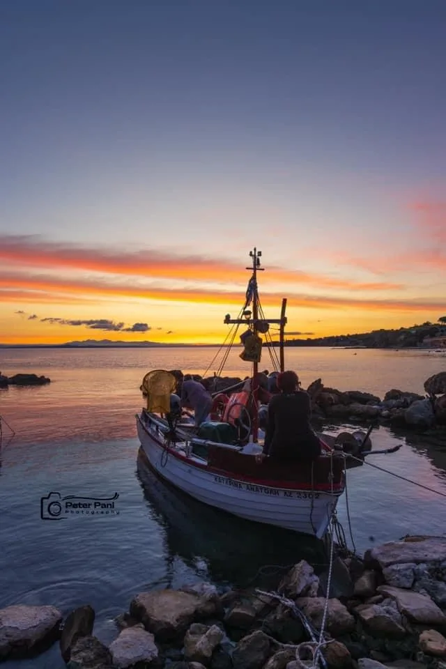 A serene sunset photograph shows a small fishing boat moored at a rocky shore, with several people aboard. The sky is…