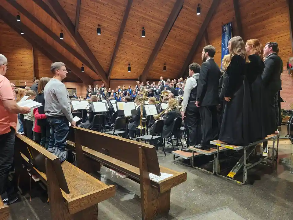 A choir and band perform in a wooden church with high vaulted ceilings. The choir members, dressed in black robes, stand on…