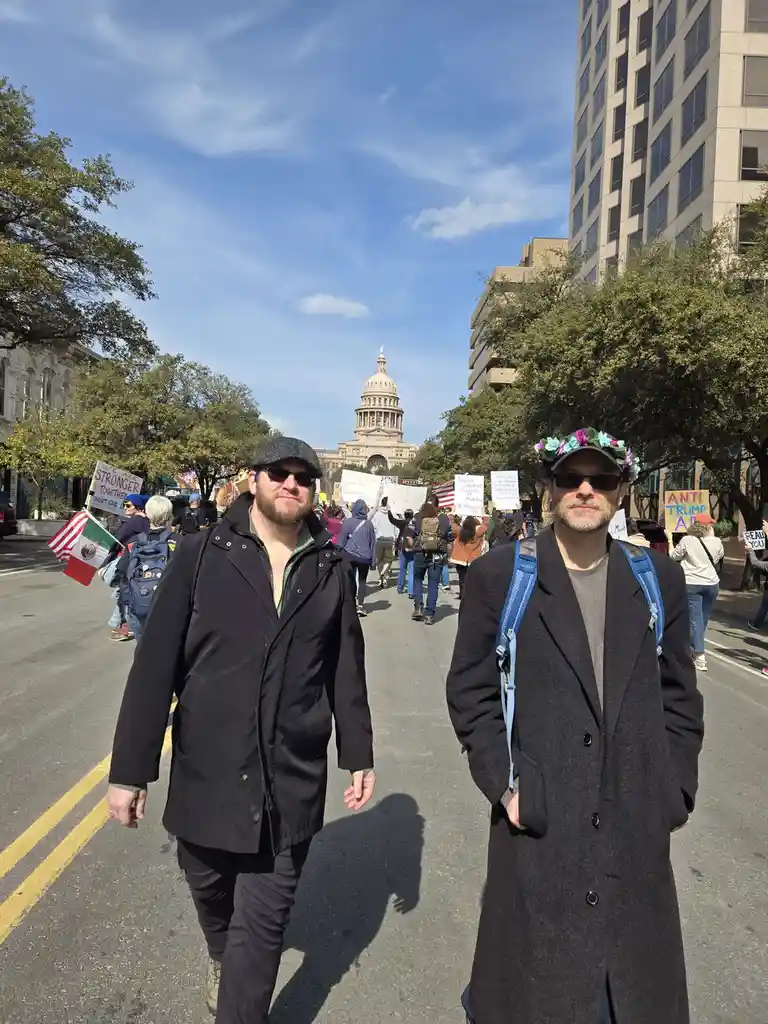 Two men walk toward the camera on a street during a protest, with the Texas State Capitol visible in the background. The man…