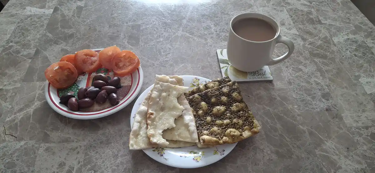 A simple meal on a marble table features a plate with sliced tomatoes and black olives, another plate with flatbread and a…
